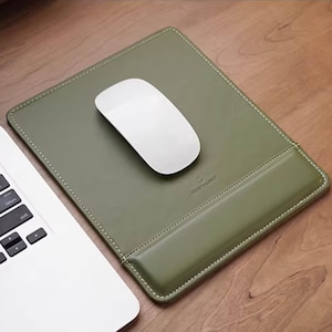 May include: Olive green leather mouse pad with a white wireless mouse. The pad has a stitched border and a wrist rest. A portion of a white laptop is visible in the corner.