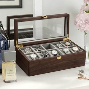 May include: A dark brown wooden watch display case with a clear glass top, showcasing twelve watches with various band styles. The interior is lined with gray fabric. Gold-colored hinges and a clasp secure the lid. The case sits on a white surface.