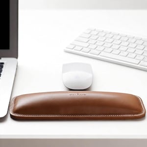 May include: A brown leather wrist rest with white stitching and the text "GRAY PLANET" is positioned in front of a white mouse and keyboard. The wrist rest is rectangular and designed for ergonomic support.