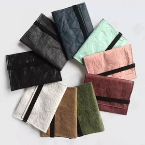 May include: A collection of colorful wallets arranged in a circular pattern. The wallets are made of textured material and feature a black elastic band closure. Colors include black, navy, mint green, pink, burgundy, white, brown, and olive green.