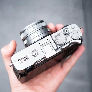 Könnte beinhalten: Silberne Fujifilm X-E5 Kamera mit Objektiv. Die Kamera verfügt über mehrere Einstellräder und Einstellungen, darunter einen Ein-/Ausschalter und Blendenwerte. Das Objektiv hat Markierungen für verschiedene Brennweiten. Die Kamera wird in einer Hand gehalten.