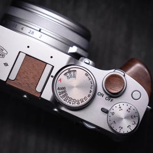 May include: Silver digital camera with a lens and wooden accents. The top of the camera features dials for settings, including shutter speed and exposure compensation. The camera has a sleek, modern design.