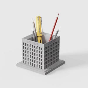 Könnte beinhalten: Ein grauer Stifthalter mit rechteckiger Basis und würfelförmiger Oberseite, mit bogenförmigen Öffnungen. Er enthält einen gelben Stift, einen roten Bleistift und graue Bleistifte. Text ist auf der Seite des Stifthalters sichtbar.