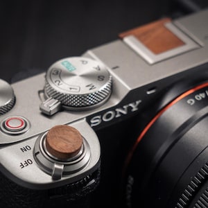 May include: Close-up of a silver Sony camera with a wooden button and dial. The lens is black with an orange accent. The camera has a textured grip and a dial with the letters P, A, S, and M. The word "SONY" is visible.