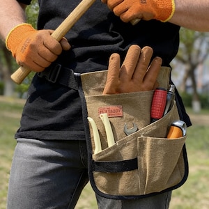 Może przedstawiać: Brązowy płócienny pas narzędziowy z wieloma kieszeniami, mieszczący narzędzia i rękawice robocze. Pas ma czarny pasek i napis "FLY DADDY" na naszywce. Osoba ma na sobie pomarańczowe rękawice robocze.