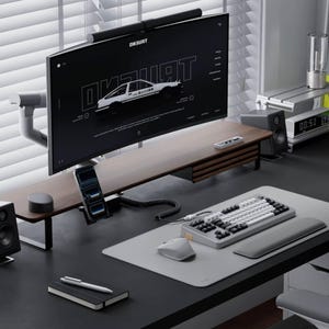 Può includere: Un moderno allestimento da scrivania con un monitor curvo che mostra una grafica. La scrivania include una tastiera, un mouse e un supporto per telefono. Sono visibili anche un piccolo altoparlante e un taccuino con una penna. La combinazione di colori è principalmente nero, marrone e grigio.