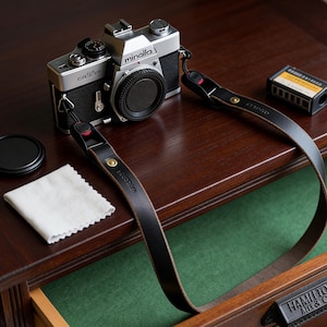 Puede incluir: Una cámara de película Minolta vintage con una correa de cuero negro y una tapa de lente se encuentra sobre una superficie de madera oscura. También hay un pequeño paño de limpieza blanco y un cartucho de película.