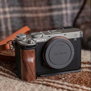 Peut inclure: Appareil photo numérique Sony Alpha 7C argenté et noir avec une poignée en bois et un bouchon d'objectif. L'appareil photo est muni d'une sangle en cuir marron. Le bouchon d'objectif porte l'inscription "SONY".