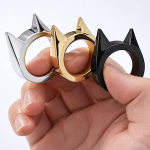 May include: Three rings in silver, gold, and black, each featuring a cat ear design. The rings are held up by a hand, showcasing the different colors and finishes. The rings are a unique fashion accessory.