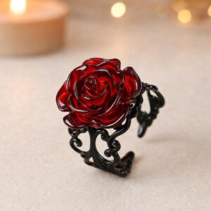 May include: A black metal cuff bracelet featuring a large, vibrant red rose. The rose is the focal point, with detailed petals. The bracelet has an open design with ornate, swirling patterns on the band.