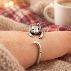 May include: A silver-colored cuff bracelet featuring a detailed mouse design. The mouse appears to be embracing the wrist. The bracelet is open-ended and adjustable. The background includes a cozy, warm-toned setting.