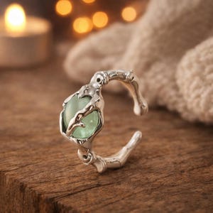 May include: A silver-toned ring with an organic, branch-like design. The ring features a light green, oval-shaped stone held in a detailed setting. The ring is displayed on a wooden surface, with a soft, blurred background.