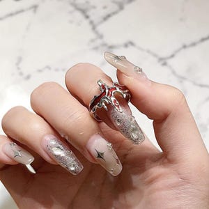 May include: A silver ring with a red flame design. The ring is worn on a finger with long, glittery, and embellished nails. The nails are painted with a clear base and adorned with silver glitter, star and diamond accents.