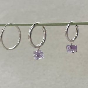 Può includere: Tre orecchini a cerchio in argento appesi a un sottile stelo verde. Il primo è un cerchio semplice. Il secondo e il terzo cerchio presentano ciascuno un piccolo ciondolo in pietra traslucida color viola chiaro di forma quadrata.