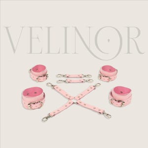 May include: Pink leather restraints set with silver-tone hardware. The set includes wrist cuffs, ankle cuffs, and connecting straps. The word "VELINOR" is displayed in a light gray font at the top of the image.