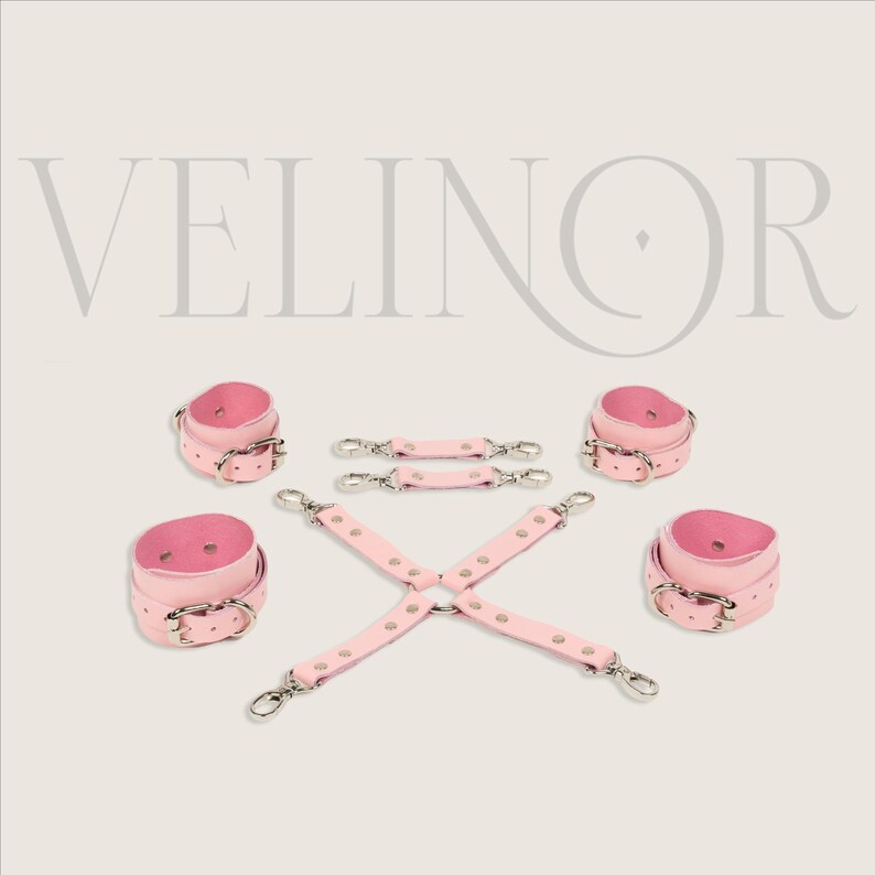 May include: Pink leather restraints set with silver-tone hardware. The set includes wrist cuffs, ankle cuffs, and connecting straps. The word "VELINOR" is displayed in a light gray font at the top of the image.