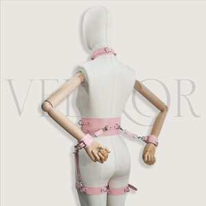 May include: A pink leather bondage set, comprising a collar, wrist cuffs, a waist belt, and leg restraints. The set is displayed on a mannequin. The metal hardware is silver-coloured.