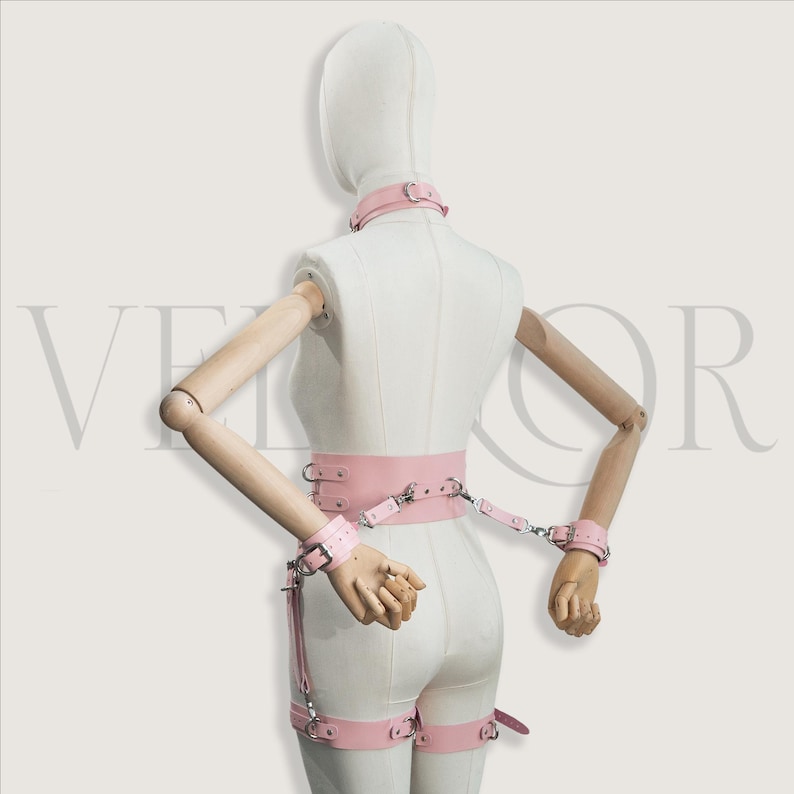 May include: A pink leather bondage set, comprising a collar, wrist cuffs, a waist belt, and leg restraints. The set is displayed on a mannequin. The metal hardware is silver-coloured.