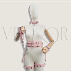 May include: A pink leather bondage set displayed on a mannequin. The set includes a choker, harness, wrist cuffs, and leg straps, all with silver-tone hardware. A leash with a handle is also included. The set is designed for role play.