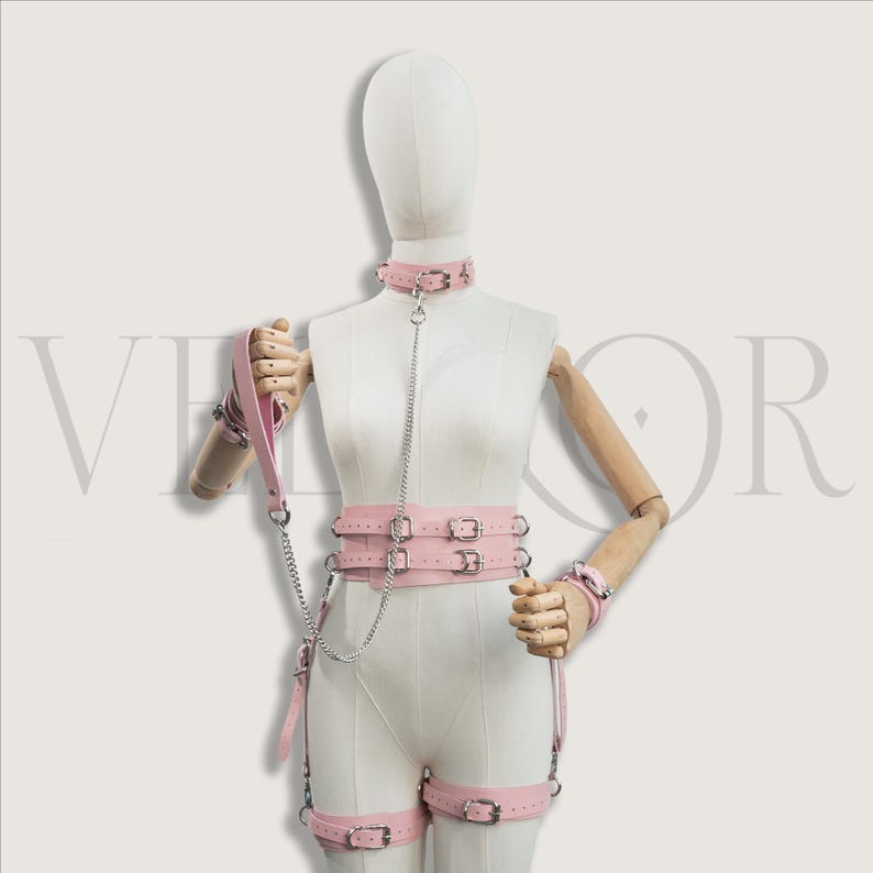 May include: A pink leather bondage set displayed on a mannequin. The set includes a choker, harness, wrist cuffs, and leg straps, all with silver-tone hardware. A leash with a handle is also included. The set is designed for role play.