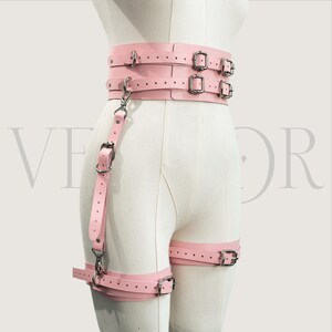 May include: A light pink leather harness with multiple straps and silver-coloured metal buckles. The harness includes a waist belt, leg straps, and a connecting strap with a clasp. The design is visible on a mannequin.