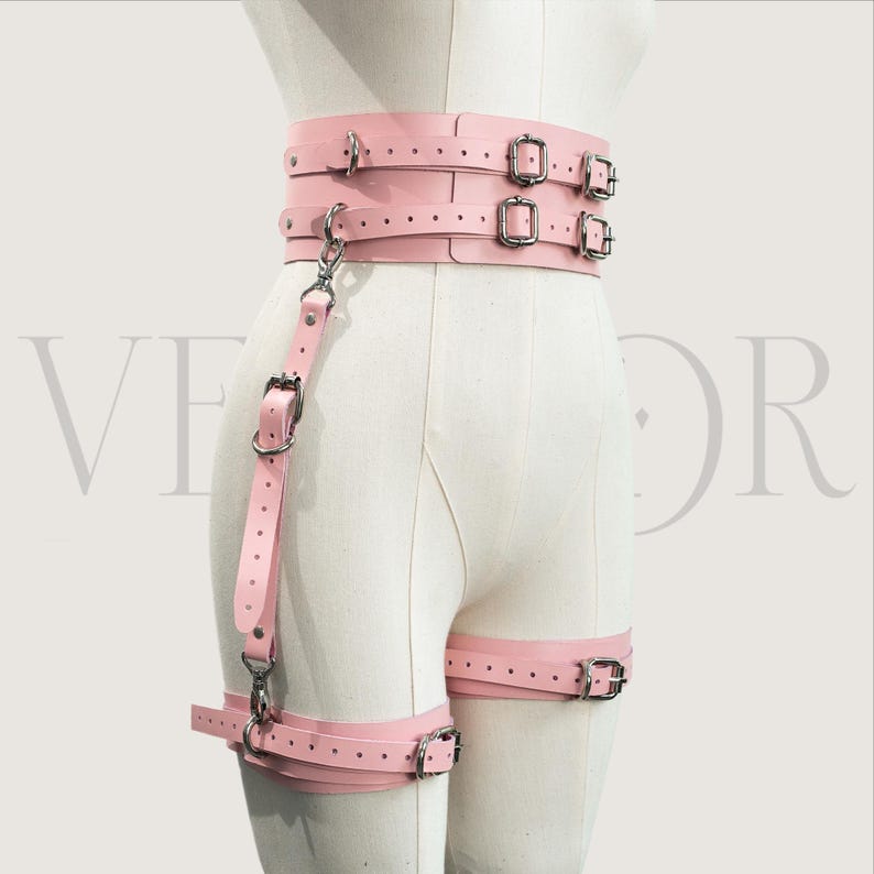 May include: A light pink leather harness with multiple straps and silver-coloured metal buckles. The harness includes a waist belt, leg straps, and a connecting strap with a clasp. The design is visible on a mannequin.