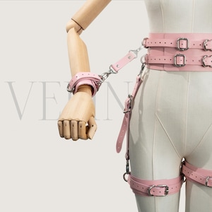 May include: A pink leather restraint set with a wrist cuff, waist belt, and leg straps. The set includes metal buckles and chain connectors. The items are displayed on a mannequin.