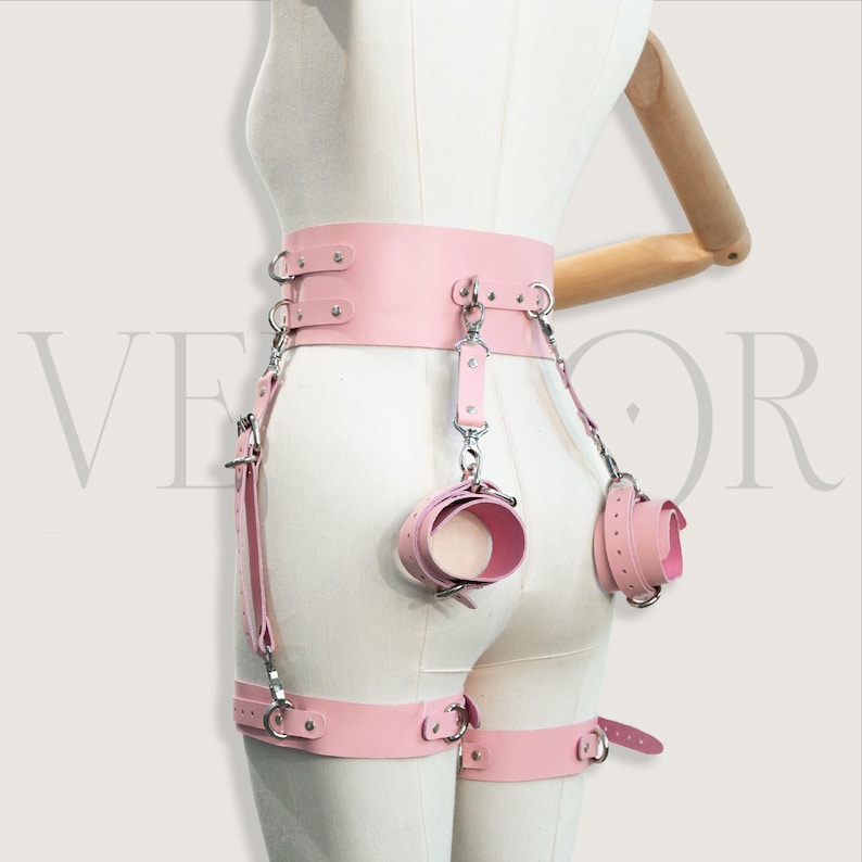 May include: A pink leather restraint harness with cuffs and leg straps. The harness has silver-tone metal hardware, including rings and clasps. Cuffs and leg straps are connected to the waist belt with chains and straps.