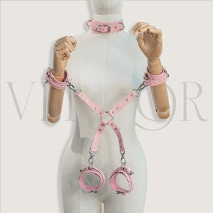 May include: A pink leather restraint set, including a collar, wrist cuffs, and ankle cuffs, connected by straps and metal hardware. The set is displayed on a mannequin. The design features a harness-style construction with adjustable buckles.