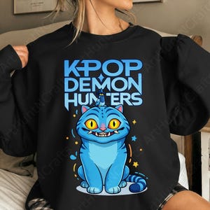 May include: Black sweatshirt featuring a blue cartoon cat with yellow eyes and the text "KPOP DEMON HUNTERS" in blue. The cat has a small blue creature on its head. The design includes stars and water droplets.