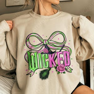 May include: Beige sweatshirt with a graphic design featuring the word "WICKED" in green and pink, a witch hat, a broom, and pink roses. The design includes a large bow with green and pink stripes, and small pink stars.