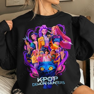 May include: Black sweatshirt with a colourful graphic of anime-style characters wielding weapons, framed by purple swirls. The text "KPOP DEMON HUNTERS" is below a blue cat with yellow eyes. The design is detailed and vibrant, suitable for fans of the genre.