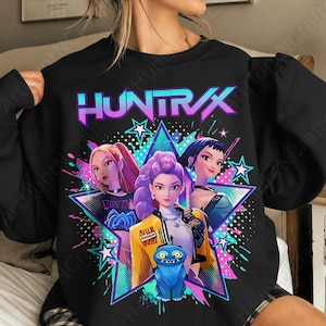 May include: Black sweatshirt featuring a graphic design with the word "HUNTRAX" in purple and blue. The design includes three stylized female figures, a blue creature, and a star-shaped background with neon accents.