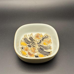 Può includere: Una ciotola quadrata in ceramica bianco sporco con un motivo floreale decorativo al centro. Il design presenta elementi neri, gialli e marroni. La ciotola ha una forma leggermente arrotondata e una superficie liscia.