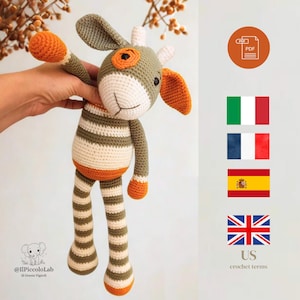 Può includere: Un giocattolo a forma di giraffa all'uncinetto fatto a mano nei toni del verde, arancione e bianco. La giraffa ha gambe e corpo a righe, con dettagli arancioni sulle orecchie, sulle braccia e sui piedi. Il giocattolo è tenuto da una mano.