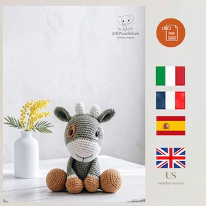 Può includere: Una giraffa all'uncinetto fatta a mano nei toni del grigio, bianco e marrone. Il peluche ha un'espressione amichevole ed è seduto. L'immagine include anche un vaso bianco con fiori gialli e un'icona PDF.