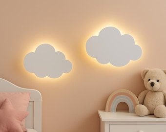 Op de afbeelding: Twee witte, wolkvormige wandlampen met warme achtergrondverlichting gemonteerd op een beige muur. Daaronder staat op een wit nachtkastje een kleine houten regenboog en een lichtbruine teddybeer naast een stervormig kussen.