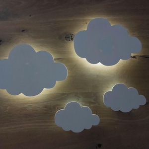 Könnte beinhalten: Vier weiße, wolkenförmige Wandleuchten sind auf einer Holzoberfläche montiert. Die Leuchten strahlen ein warmes Leuchten hinter den Wolkenausschnitten aus und erzeugen einen weichen, umgebenden Lichteffekt. Die Wolken sind unterschiedlich groß.