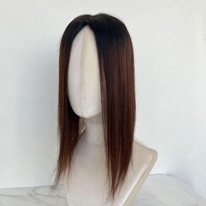 以下が含まれることがあります： マネキンの頭に展示されている茶色と黒のヘアートッパー。髪はストレートで肩まで届きます。ヘアートッパーは、濃い茶色の根元から明るい茶色にフェードしています。