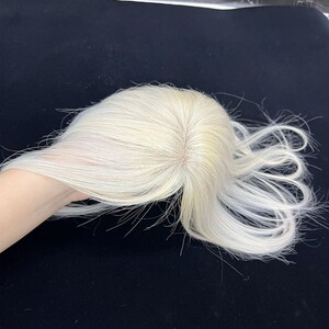 May include: A light blonde hair topper with a natural-looking part. The hair is styled with loose waves and is held in a hand against a dark background. The hairpiece appears to be made of human hair.