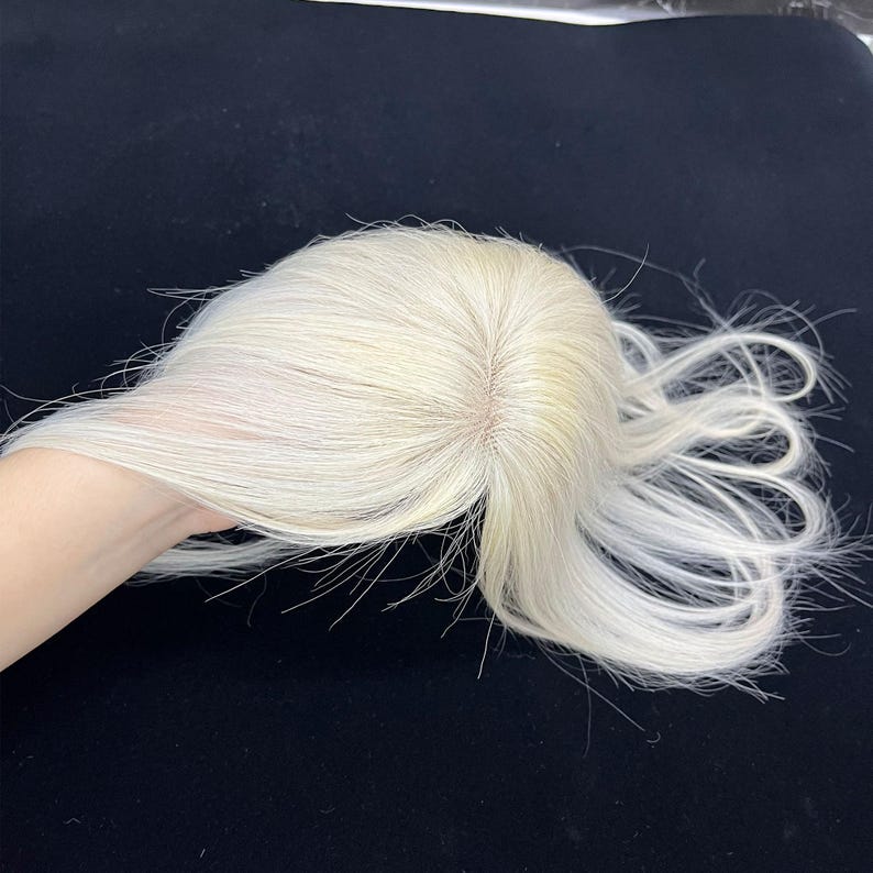 May include: A light blonde hair topper with a natural-looking part. The hair is styled with loose waves and is held in a hand against a dark background. The hairpiece appears to be made of human hair.