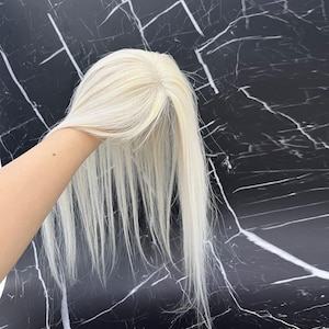 Könnte beinhalten: Ein hellblonder Haaraufsatz mit langen, geraden Strähnen. Das Haarteil wird vor einem schwarz-weißen Marmorhintergrund gehalten. Das Haarteil scheint aus Kunstfasern zu bestehen.