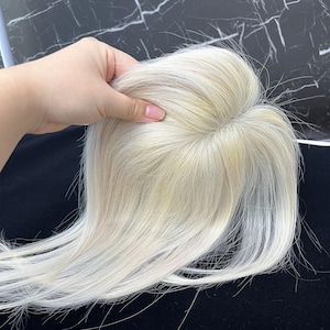 May include: A light blonde hair topper, held by a hand, against a dark background. The hairpiece appears to be made of real hair, with a natural look and feel. The hair is styled with a slight wave.