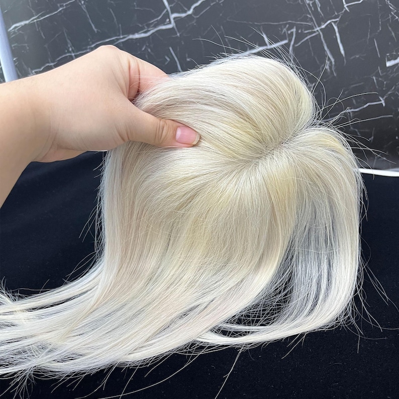 May include: A light blonde hair topper, held by a hand, against a dark background. The hairpiece appears to be made of real hair, with a natural look and feel. The hair is styled with a slight wave.