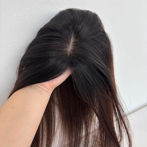 以下が含まれることがあります： 長いストレートヘアのダークブラウンのヘアピース。ヘアピースは持ち上げられ、自然に見える分け目と髪の質感を披露しています。髪はわずかな光沢のある単色に見えます。