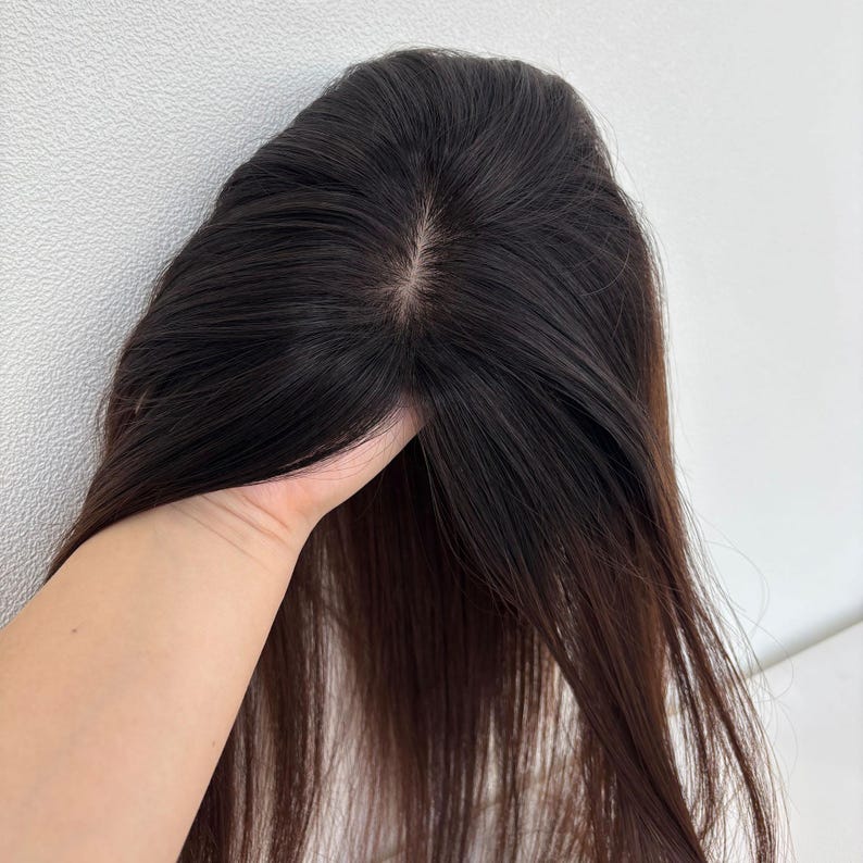 以下が含まれることがあります： 長いストレートヘアのダークブラウンのヘアピース。ヘアピースは持ち上げられ、自然に見える分け目と髪の質感を披露しています。髪はわずかな光沢のある単色に見えます。