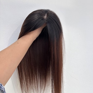 以下が含まれることがあります： 長いストレートヘアのダークブラウンのヘアピース。ヘアピースは手で持たれており、暗い根元から明るい毛先への自然な色のグラデーションを示しています。ヘアピースはボリュームと長さを加えるように設計されています。