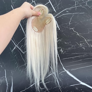 May include: A blonde hair topper with three clips attached to a small, skin-toned base. The hair is long and straight, with a natural-looking texture. The topper is designed to add volume and length to existing hair.