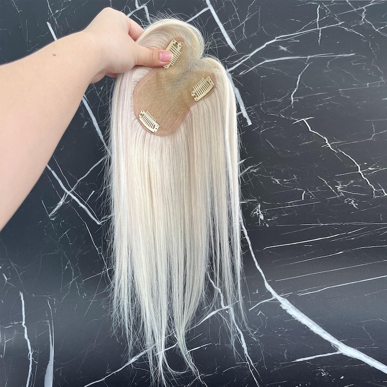 May include: A blonde hair topper with three clips attached to a small, skin-toned base. The hair is long and straight, with a natural-looking texture. The topper is designed to add volume and length to existing hair.