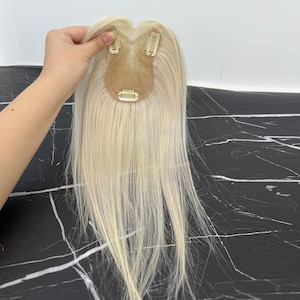 May include: A blonde hair topper with three small clips attached. The hairpiece is a light, natural blonde color and appears to be made of real hair. The clips are a light gold color.